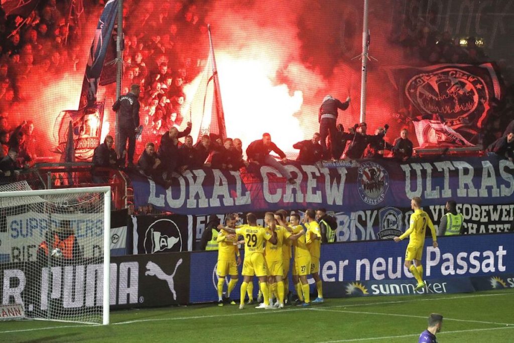 Die Mannschaft von Arminia Bielefeld jubelt vor dem eigenen Fanblock, die Fans von Arminia Bielefeld haben Bengalos gezündet; Foto: Imago images / Joachim Sielski