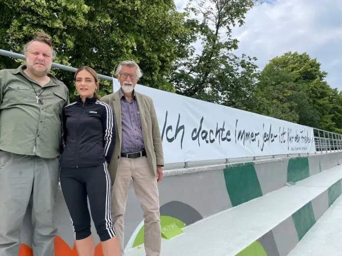 (von links) Künstler Volker-Johannes Trieb, Moskaubadleiterin Kerstin Klotz und Reinhold Mokrosch von der Remarque-Gesellschaft vor dem Banner auf der Tribüne des Moskaubads. / Foto: Stadtwerke Osnabrück (von links) Künstler Volker-Johannes Trieb, Moskaubadleiterin Kerstin Klotz und Reinhold Mokrosch von der Remarque-Gesellschaft vor dem Banner auf der Tribüne des Moskaubads. / Foto: Stadtwerke Osnabrück